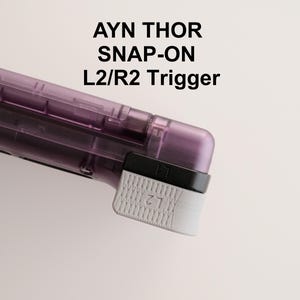 Könnte beinhalten: Ein lila-schwarzer Snap-On L2/R2-Trigger für das Handheld-Gaming-Gerät AYN THOR. Der Trigger hat eine weiße, strukturierte Oberfläche mit den Buchstaben "L2" aufgedruckt. Der Text "AYN THOR SNAP-ON L2/R2 Trigger" ist auf dem Bild aufgedruckt.