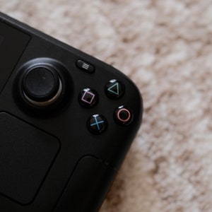 May include: Black handheld gaming console with a joystick, buttons, and a directional pad. The buttons are colored pink, green, blue, and red. The console is on a neutral-colored, textured surface.