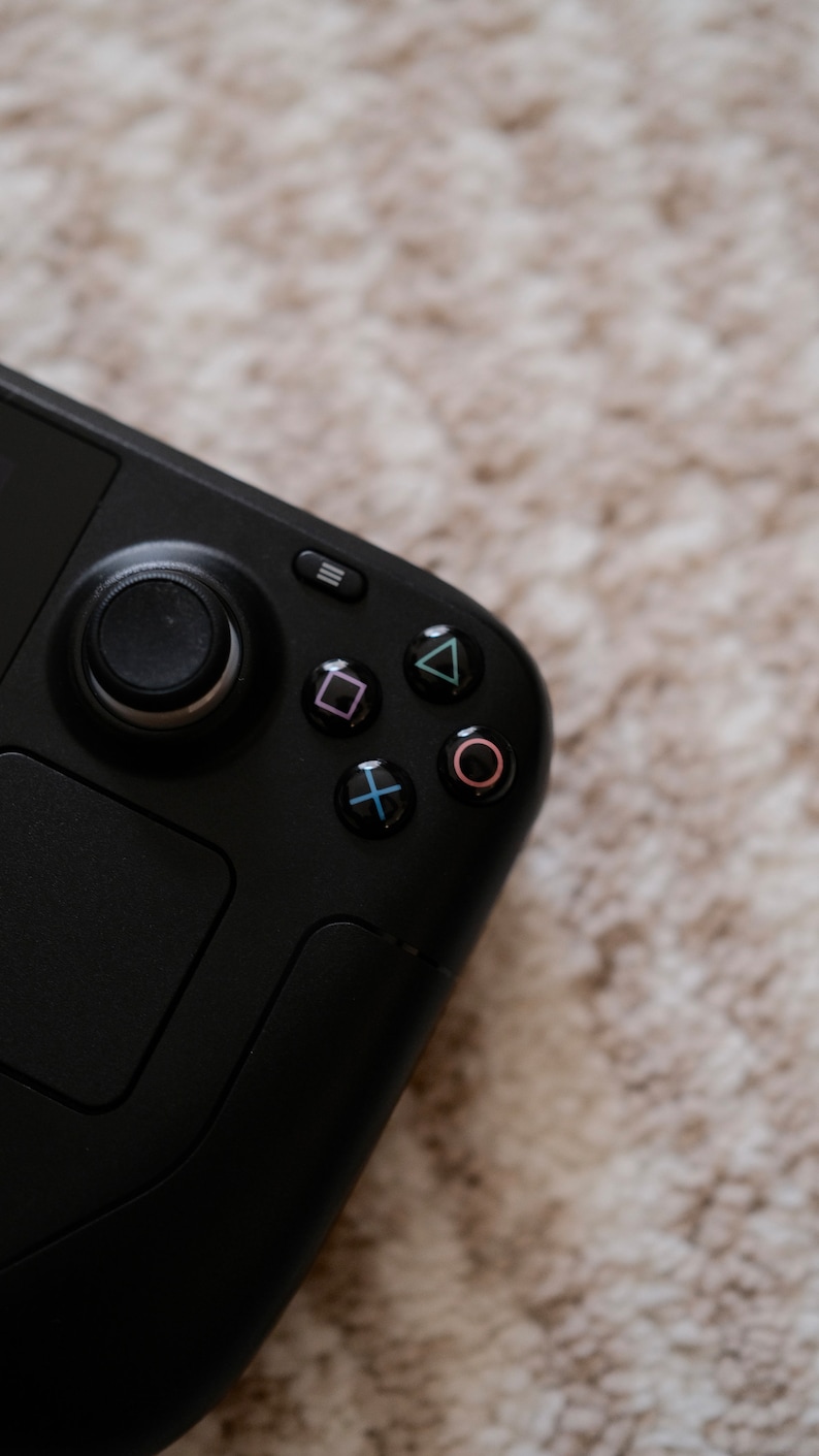 May include: Black handheld gaming console with a joystick, buttons, and a directional pad. The buttons are colored pink, green, blue, and red. The console is on a neutral-colored, textured surface.