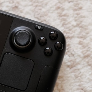 Puede incluir: Un dispositivo de juego portátil negro con un botón circular y cuatro botones más pequeños en el lado derecho. El dispositivo tiene una pantalla táctil y un panel táctil negro.