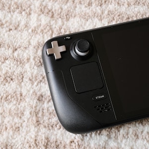 May include: Black handheld gaming device with a screen, buttons, and a joystick. The device has a directional pad, a thumbstick, and a touchpad. The word "STEAM" is printed on the device. The device is on a beige and white patterned surface.