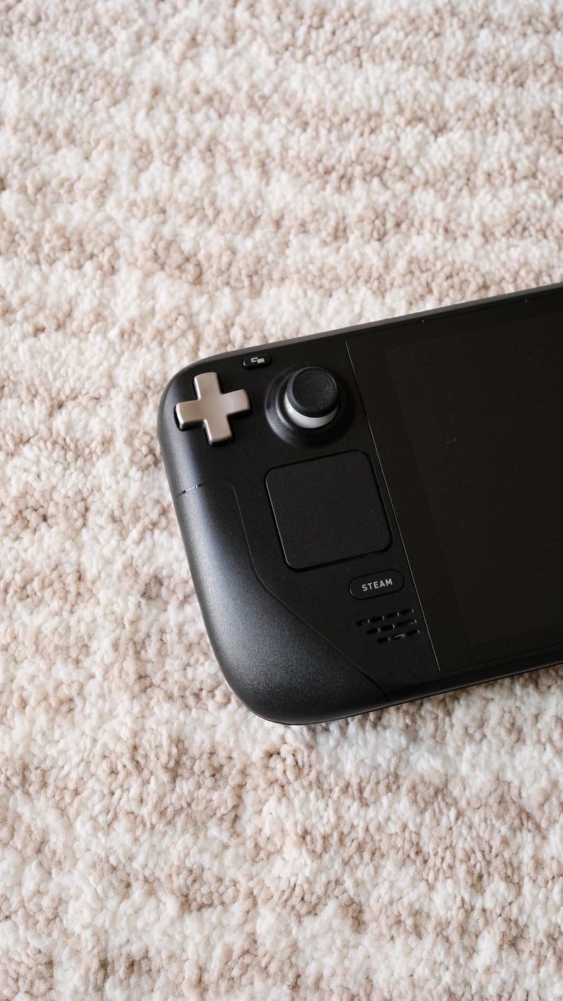 May include: Black handheld gaming device with a screen, buttons, and a joystick. The device has a directional pad, a thumbstick, and a touchpad. The word "STEAM" is printed on the device. The device is on a beige and white patterned surface.