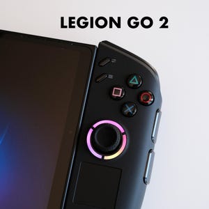 以下が含まれることがあります： 上部に「LEGION GO 2」の文字が入った黒い携帯型ゲーム機のクローズアップ。デバイスは左側に画面、右側にコントローラーがあり、カラフルなボタンと虹色のライトが付いたジョイスティックを備えています。