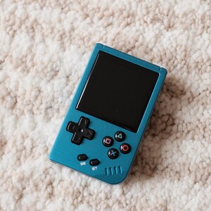May include: A teal handheld gaming console with a black screen and black control buttons. The console features a directional pad, action buttons, and start/select buttons. The device is resting on a cream-colored, textured surface.
