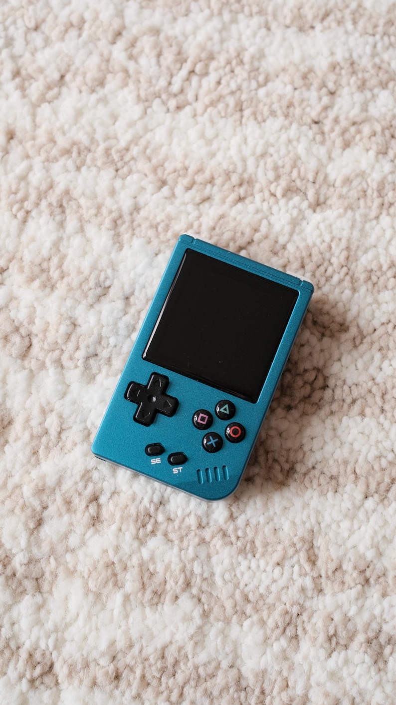 May include: A teal handheld gaming console with a black screen and black control buttons. The console features a directional pad, action buttons, and start/select buttons. The device is resting on a cream-colored, textured surface.