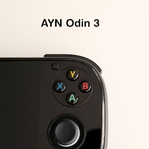 Peut inclure: Gros plan sur une console de jeu portable noire avec le texte "AYN Odin 3" en haut. L'appareil comprend des boutons d'action colorés (Y, X, B, A), une croix directionnelle et un joystick. Le fond est blanc cassé.