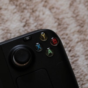 Puede incluir: Un mando de juego negro con cuatro botones etiquetados como A, B, X e Y. Los botones son de color rojo, verde, azul y amarillo.