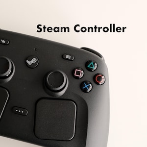 Op de afbeelding: Een zwarte Steam Controller videogame-gamepad met twee analoge sticks, een centrale Steam-logoknop, een menuknop en vier actieknoppen met geometrische symbolen in blauw, rood en roze. Twee grote vierkante touchpads bevinden zich aan de onderkant.