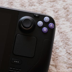 May include: Close-up of a black handheld gaming device. The device features a black joystick, a directional pad, and purple buttons labeled A, B, X, and Y. The device is on a light-colored, textured surface.