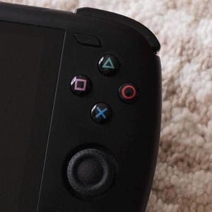 May include: A black handheld gaming device with a directional pad and four action buttons. The buttons are labeled with the symbols: triangle, square, circle, and cross.