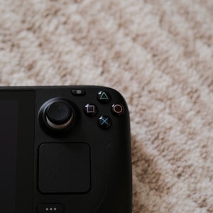 May include: Close-up of a black handheld gaming console. The device features a screen, a joystick, a touchpad, and several buttons, including a directional pad and action buttons. The background is a light-colored, textured surface.