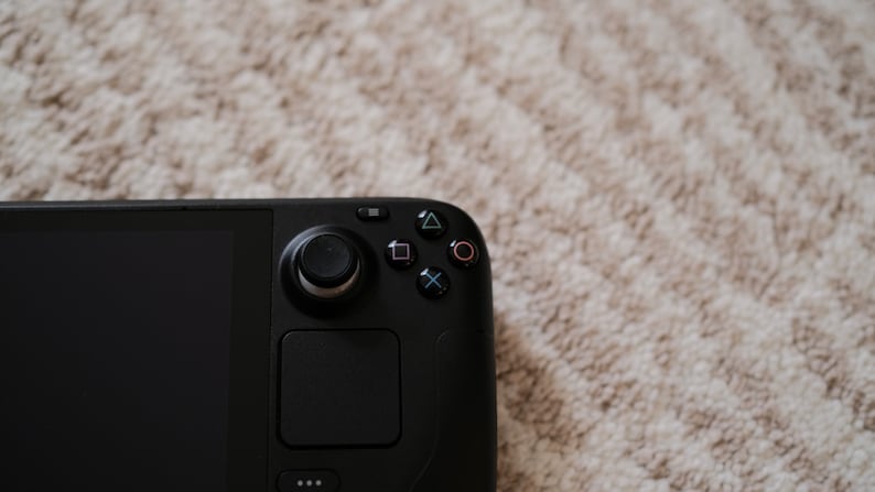 May include: Close-up of a black handheld gaming console. The device features a screen, a joystick, a touchpad, and several buttons, including a directional pad and action buttons. The background is a light-colored, textured surface.