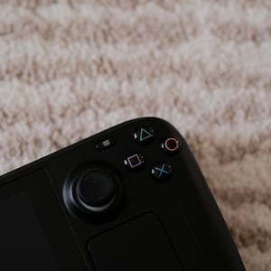 May include: A black handheld gaming console with a joystick, buttons, and a touchpad. The buttons include a triangle, square, X, and circle. The console is on a textured, light-colored surface.