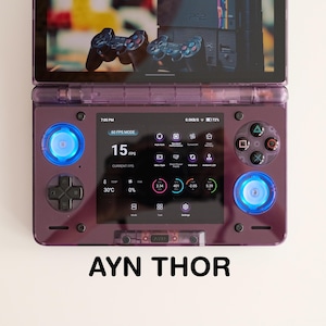 May include: A purple handheld gaming console with a screen displaying game information. The console has blue illuminated buttons and the text "AYN THOR" at the bottom. The screen shows the FPS at 15.