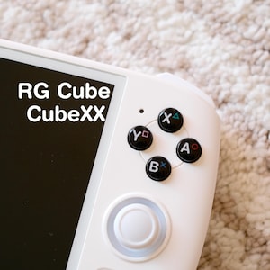 May include: A white handheld games console featuring black buttons marked X, Y, A, and B. The console has a black screen and the text "RG Cube CubeXX" is displayed. The device rests on a textured, light-coloured surface.