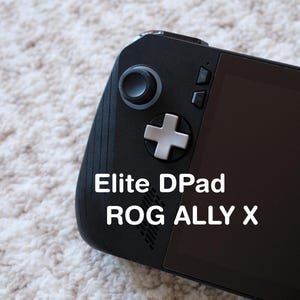 May include: A black handheld gaming device with the text "Elite DPad ROG ALLY X" in white. The device features a D-pad, buttons, and a screen. The background is a light-colored, textured surface.