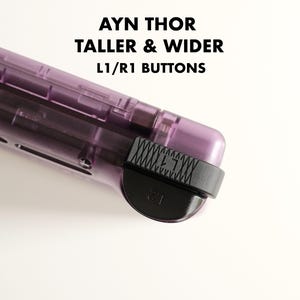 Op de afbeelding: Een close-up van een paars draagbaar gamingapparaat met zwarte L1/R1-knoppen. De tekst "AYN THOR TALLER & WIDER L1/R1 BUTTONS" staat in het zwart bovenaan de afbeelding.