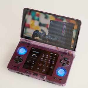 May include: A transparent purple handheld gaming console with a screen displaying a game interface. The console features a D-pad, buttons, and glowing blue lights. The screen shows a PlayStation console and controllers.
