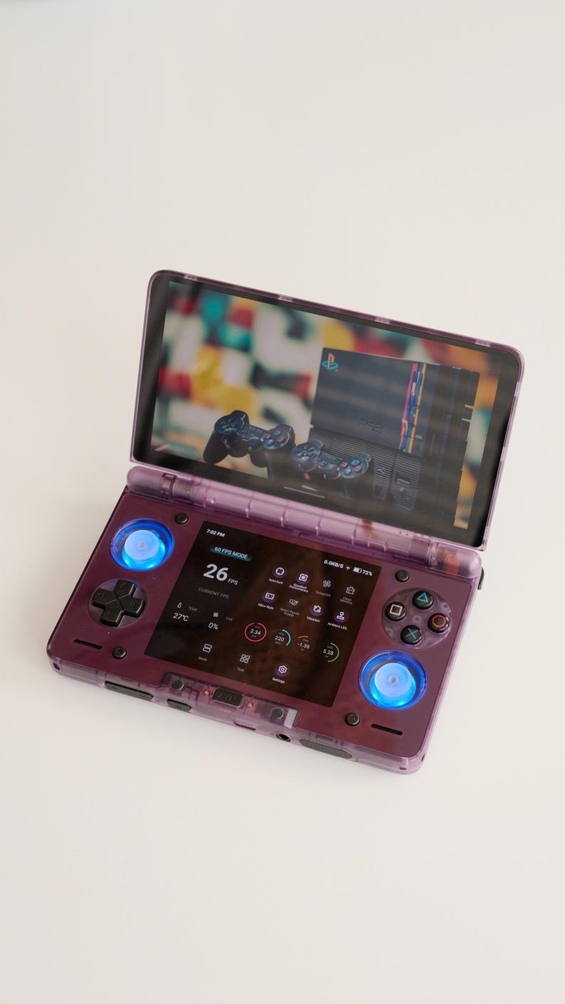 May include: A transparent purple handheld gaming console with a screen displaying a game interface. The console features a D-pad, buttons, and glowing blue lights. The screen shows a PlayStation console and controllers.