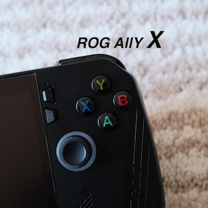 May include: A close-up of a black handheld gaming console with the text "ROG Ally X". It has a screen, a directional pad, and coloured action buttons (Y, X, B, A). The background is a light-coloured, textured surface.