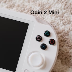 May include: White handheld gaming console featuring a black screen, a white joystick, and four coloured buttons: green triangle, pink square, red circle, and blue X. The text "Odin 2 Mini" is visible.
