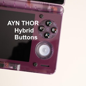 May include: A close-up of a purple handheld gaming device with black buttons and a screen. The text "AYN THOR Hybrid Buttons" is displayed on the screen. The device has a transparent purple casing.
