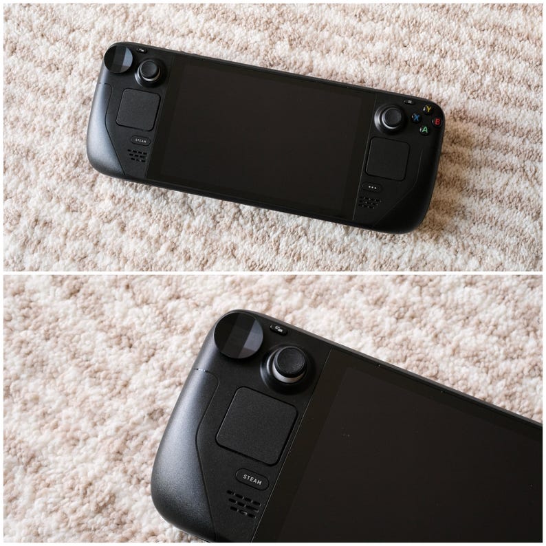 May include: A black handheld gaming console with a large screen, two thumbsticks, and various buttons. The console has a sleek, modern design and features the "STEAM" logo. The device is shown on a light-colored, textured surface.