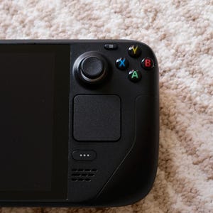 May include: A black handheld gaming console with a large screen and integrated controls. The device features a joystick, buttons, and a touchpad on the right side. The console is resting on a textured, light-colored surface.
