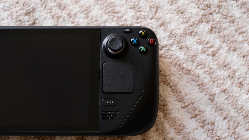 May include: A black handheld gaming console with a large screen and integrated controls. The device features a joystick, buttons, and a touchpad on the right side. The console is resting on a textured, light-colored surface.