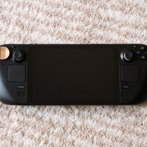 May include: A black handheld gaming console with a large screen, two control pads, and buttons. The console has a sleek design and is placed on a textured, light-colored surface.