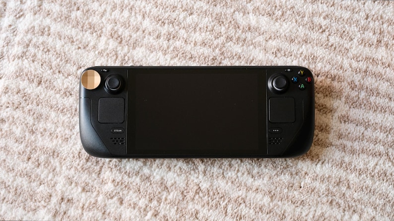 May include: A black handheld gaming console with a large screen, two control pads, and buttons. The console has a sleek design and is placed on a textured, light-colored surface.