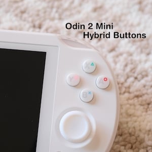 May include: Close-up of a white handheld gaming device with the text "Odin 2 Mini Hybrid Buttons." The device features a black screen and several colored buttons, including a directional pad and action buttons.