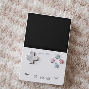 May include: A white handheld gaming console with a black screen, a white directional pad, and four white buttons with coloured symbols. The console has the text "Analogue Pocket" printed on the front.