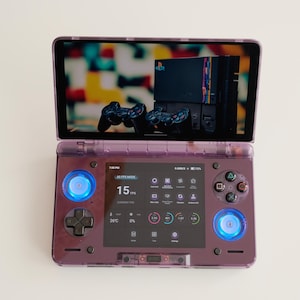 May include: A purple handheld gaming console with a screen displaying a retro gaming setup. The console features a D-pad, buttons, and two joysticks. The screen shows a PlayStation console and controllers. The console has blue illuminated lights.