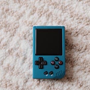 May include: A teal handheld gaming console with a black screen and colorful buttons. The device features a directional pad, action buttons, and a small speaker. The console is set against a neutral, textured background.