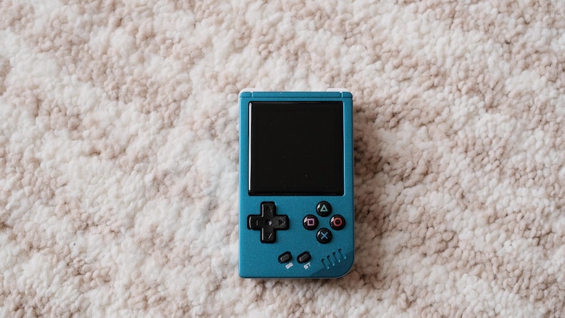 May include: A teal handheld gaming console with a black screen and colorful buttons. The device features a directional pad, action buttons, and a small speaker. The console is set against a neutral, textured background.