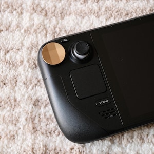 May include: A black handheld gaming device with a screen, buttons, and a trackpad. The device has a gold-colored circular button and a black joystick. The word "STEAM" is printed on the device. The device is resting on a textured, off-white surface.