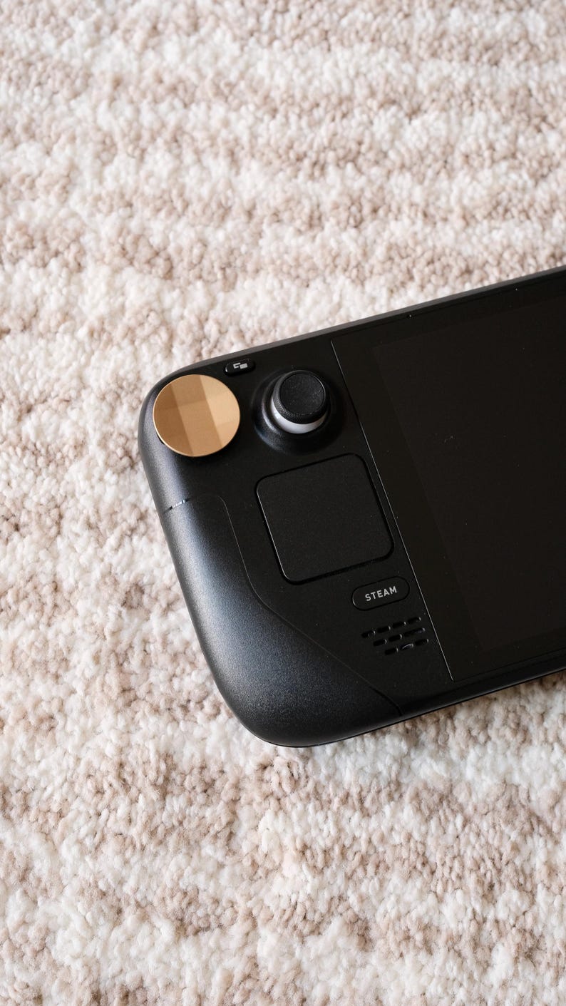 May include: A black handheld gaming device with a screen, buttons, and a trackpad. The device has a gold-colored circular button and a black joystick. The word "STEAM" is printed on the device. The device is resting on a textured, off-white surface.