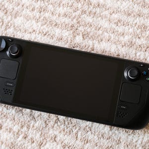 May include: A black handheld gaming console with a large screen, two control sticks, and various buttons. The device is resting on a beige, textured surface, suggesting a soft rug or carpet. The console's design is sleek and modern.