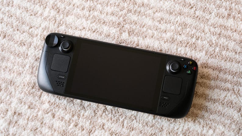 May include: A black handheld gaming console with a large screen, two control sticks, and various buttons. The device is resting on a beige, textured surface, suggesting a soft rug or carpet. The console's design is sleek and modern.