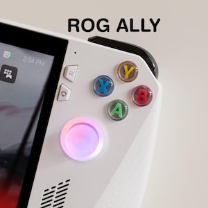 May include: A white handheld gaming device with the text "ROG ALLY" at the top. The device has colourful buttons labelled A, B, X, and Y. A glowing pink and purple light surrounds the joystick. The screen displays icons.