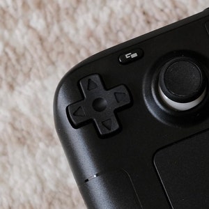 Könnte beinhalten: Ein schwarzer Handheld-Gaming-Controller mit einem Steuerkreuz, einem runden Knopf und einem Touchpad.