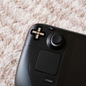 May include: Close-up of a black handheld gaming console. The console features a directional pad, a joystick, and a touch pad. The word "STEAM" is printed on the bottom of the console. The background is a light-colored, textured surface.