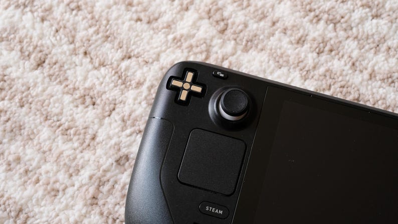 May include: Close-up of a black handheld gaming console. The console features a directional pad, a joystick, and a touch pad. The word "STEAM" is printed on the bottom of the console. The background is a light-colored, textured surface.