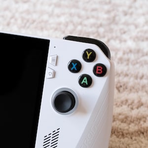 Könnte beinhalten: Eine weiße Handheld-Spielkonsole mit einem schwarzen Bildschirm und farbigen Tasten. Die Tasten sind mit den Buchstaben A, B, X und Y beschriftet. Die Konsole hat auch einen schwarzen Joystick und mehrere weiße Tasten.