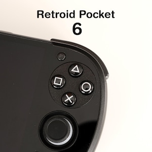 Può includere: Primo piano di un dispositivo di gioco portatile nero. Il dispositivo presenta un D-pad, pulsanti azione (triangolo, quadrato, cerchio e X) e un joystick analogico. Il testo "Retroid Pocket 6" è visualizzato in alto.