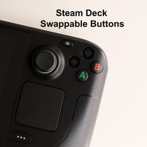 Puede incluir: Una consola de juegos portátil negra con el texto Steam Deck Swappable Buttons. El dispositivo incluye un joystick negro, un panel táctil cuadrado y cuatro botones de acción, con el botón A en verde y el botón B en rojo, además de menús.