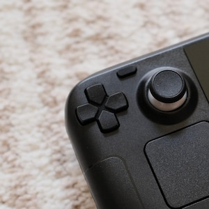 Op de afbeelding: Een zwart draagbaar gameapparaat met een ronde knop, een directionele pad en een touchpad.