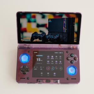 May include: A purple handheld gaming console with a screen displaying a retro gaming setup. The console features a lower screen with icons and controls, and blue illuminated buttons. The top screen shows a classic game console and controllers.