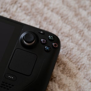 May include: A black handheld gaming device with a joystick, buttons, and a touchpad. The device has a sleek, modern design with a matte finish. The buttons are marked with colored symbols. The device is resting on a light-colored, textured surface.